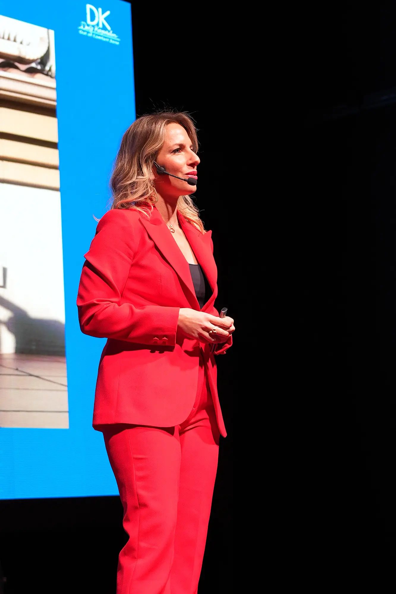 Keynote-Speakerin Deniz Kayadelen steht auf einer Bühne in einem roten Blazer gekleidet. Im Hintergrund sieht man einen Teil ihrer Präsentation.